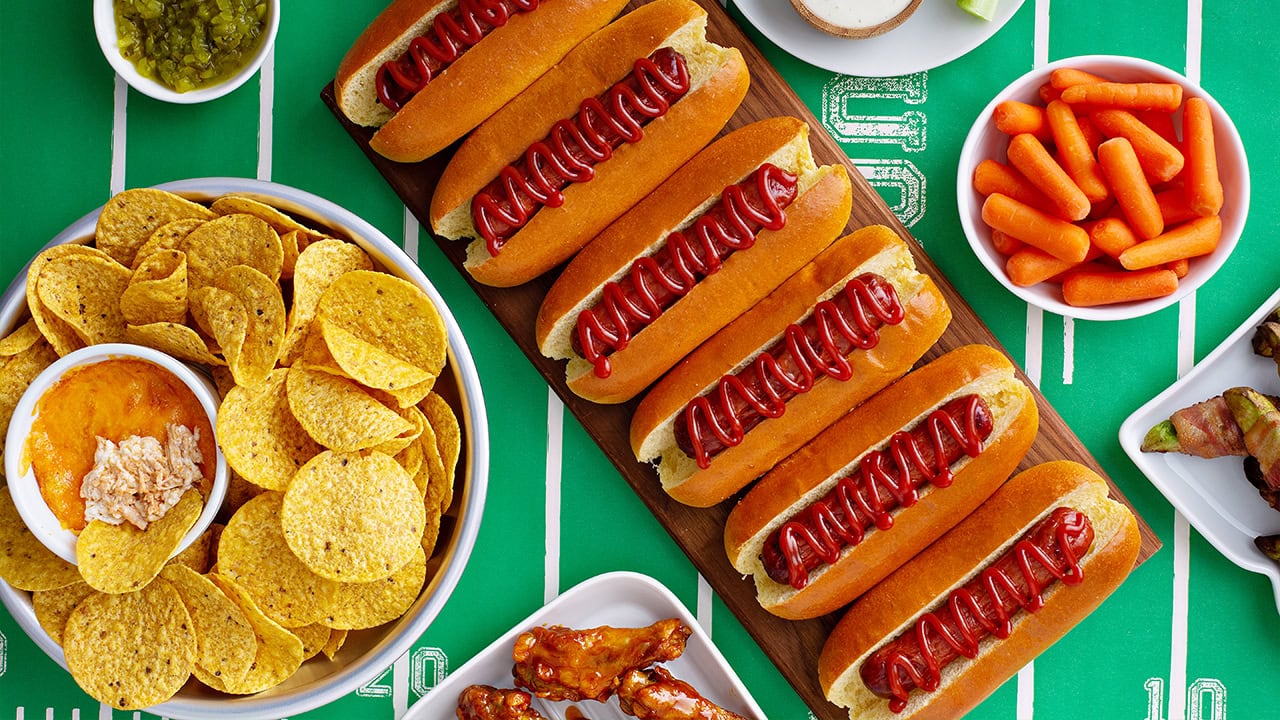 Assortment of foods like hot dogs, nachos, carrots, and wings on football field background. Image produced by More Jersey.