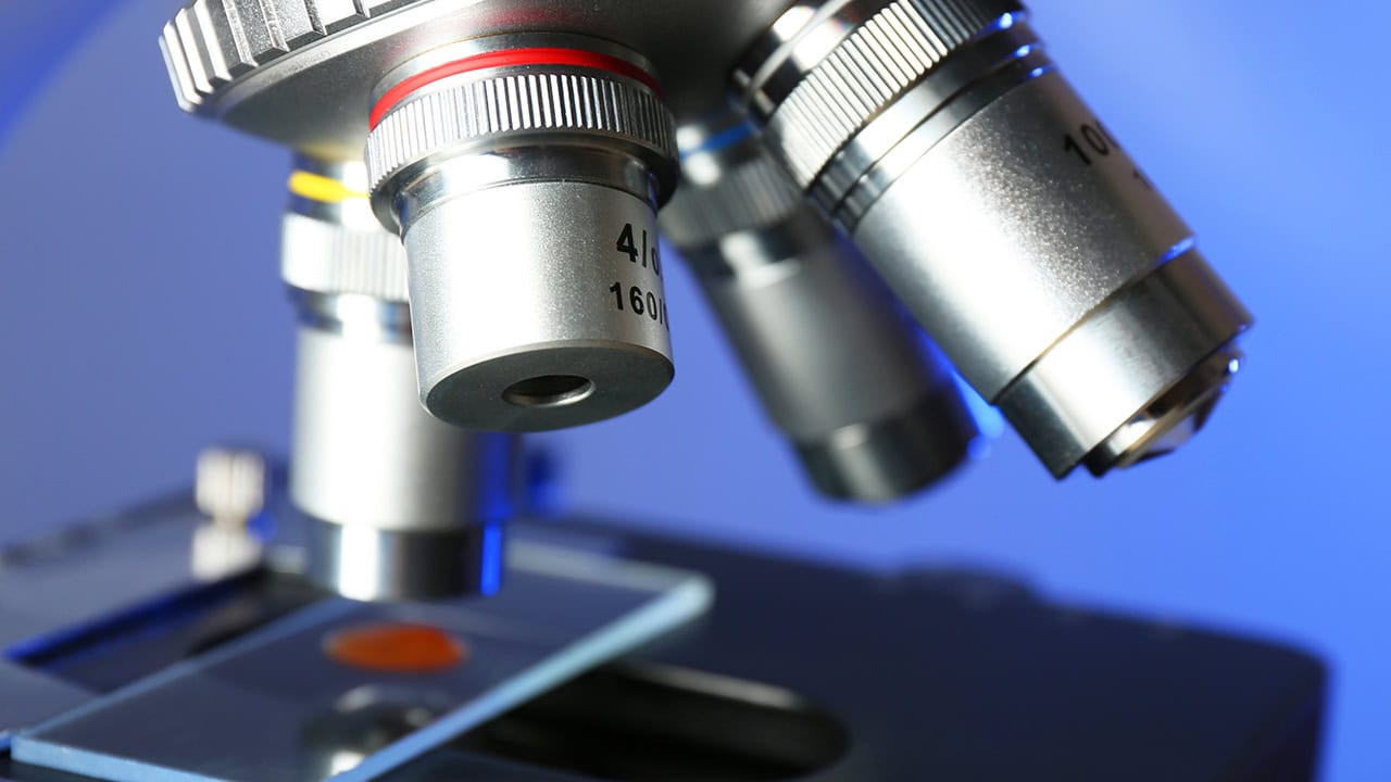 Close-up of a microscope examining a microscope slide on a blue color background. Image produced by More Jersey.