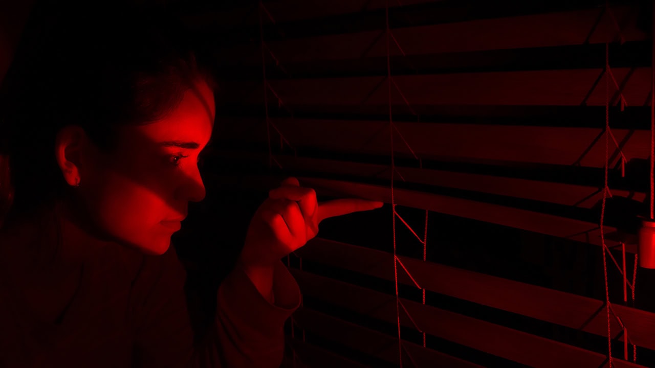 Curious woman peeking through window blinds at night with red light on. Image produced by More Jersey.