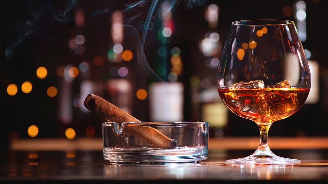 Glass of liquor with ice cubes and smoking cigar.