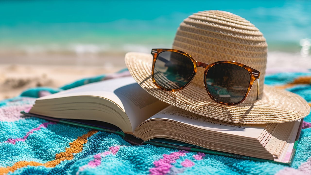 Summer hat with sunglasses on the brim over an open book on top of a towel on the beach.