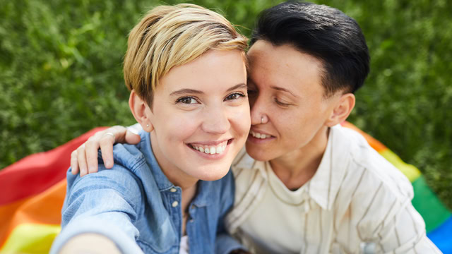 White lesbian couple taking a selfie while sitting on a rainbow flag in a local park.