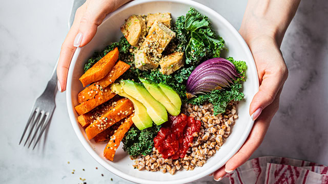 Woman holding a vegan salad of baked vegetables, avocado, and tofu.