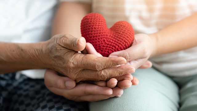 Young and senior woman holding each other's hands and a red heart made of yarn.